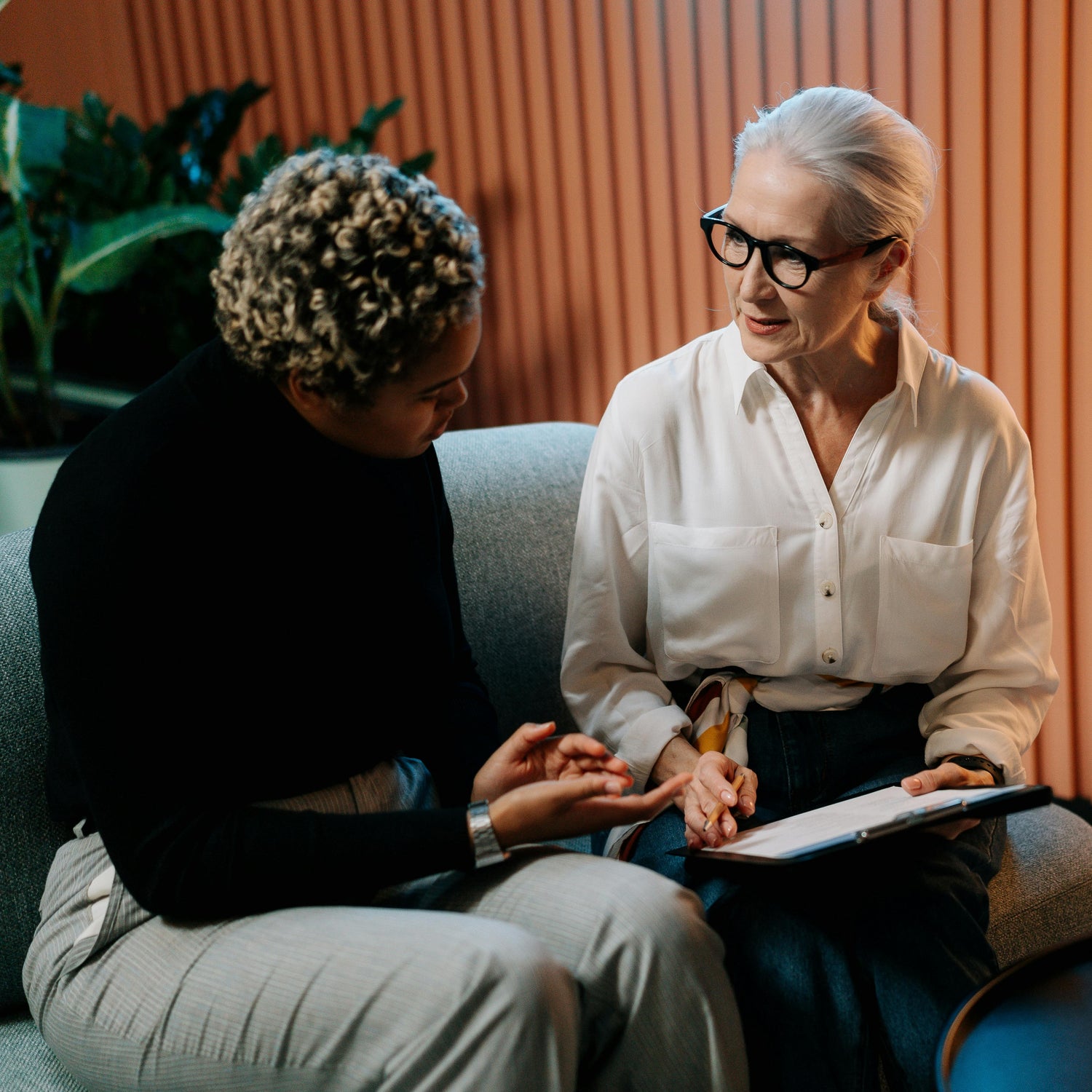 Two women sitting on a couch, one holding a tablet, in a room with plants and a lamp.